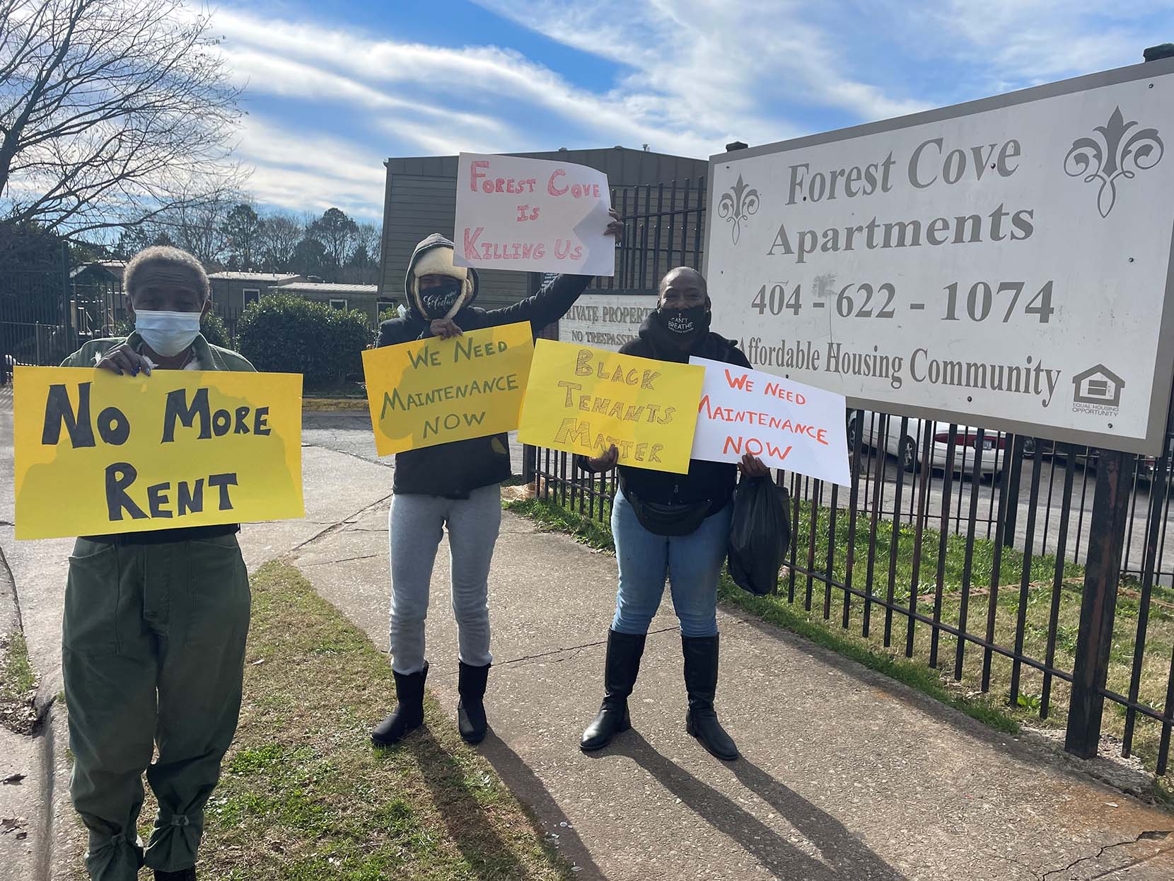 Three people standing outside a fence with signs protesting rent increases