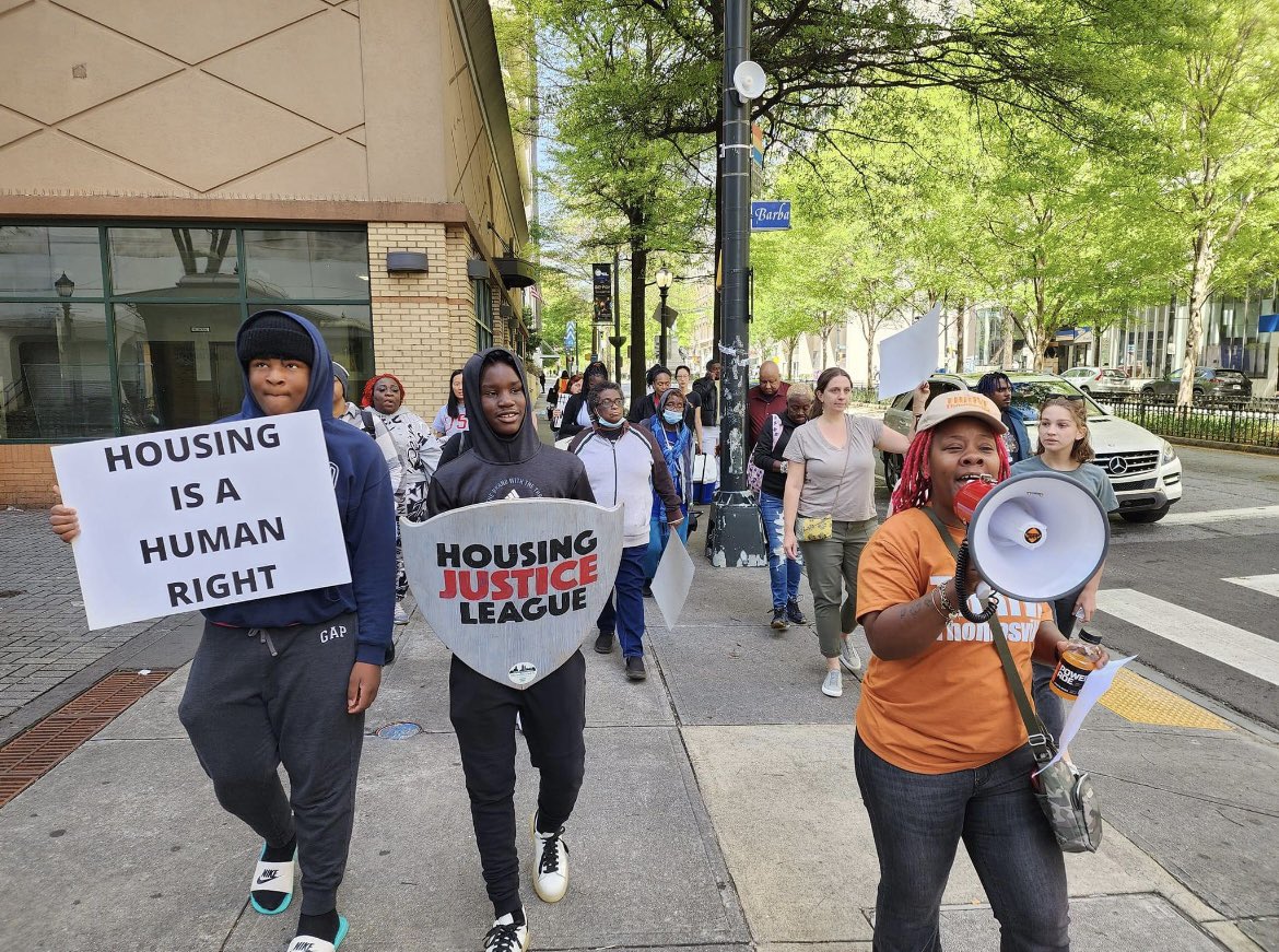 Over a dozen protesters walk on a sidewalk holding signs