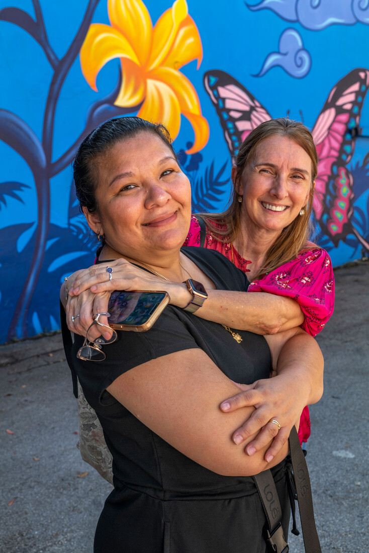 image of two people hugging and looking at camera while there is a blue mural behind them with pink butterflies