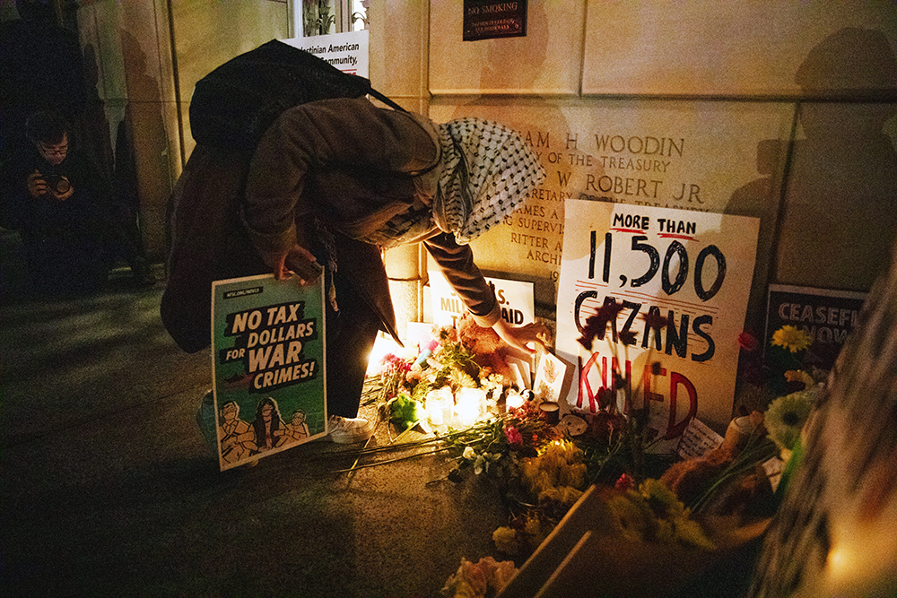 person wearing keffiyeh kneeling to put flowers at a memorial vigil