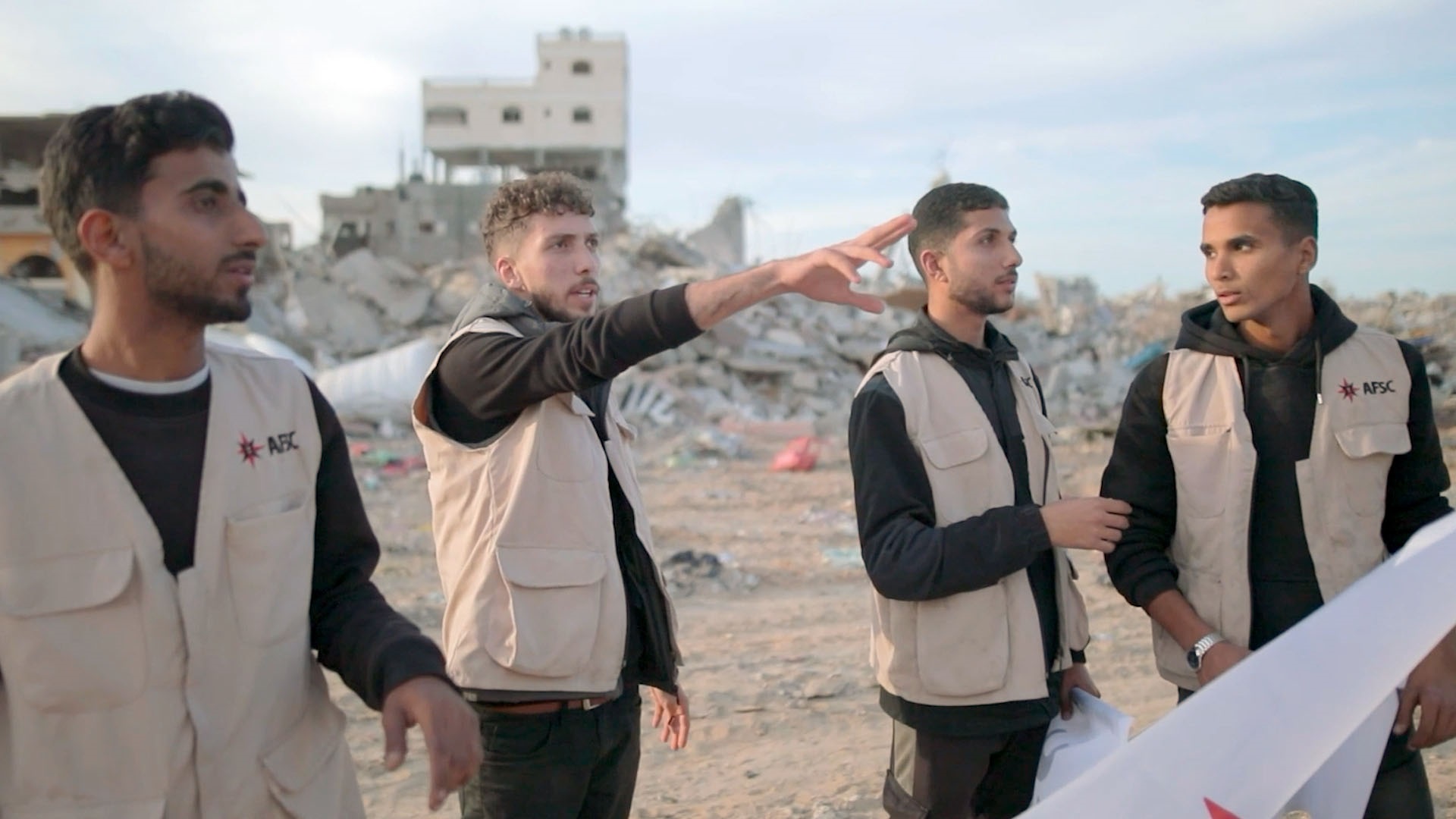 4 people in khaki jackets stand with ruins behind them