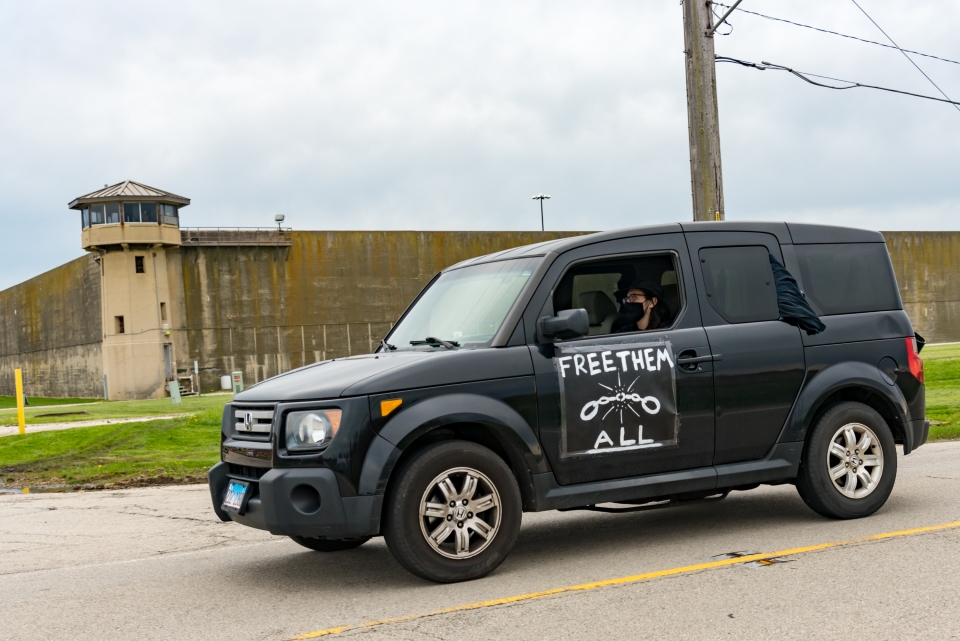 A black car drives in front of a detention center with a poster taped to the side of the car that reads "Free Them All."