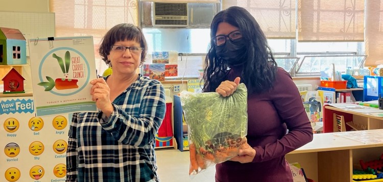 Two women pose for the camera. One holds a poster, the other a bag of carrots.